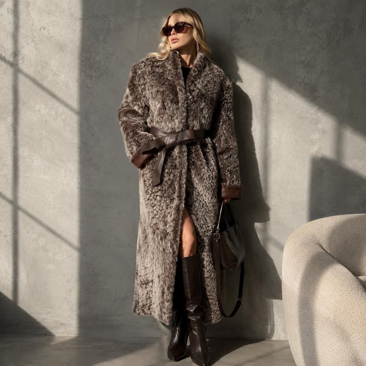 Floor-length frost-tipped reversible shearling coat with a leather belt, against a sunlit concrete wall